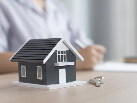 Image of a miniature house on a desk, with a blurred background of a person writing on a blank parchment.